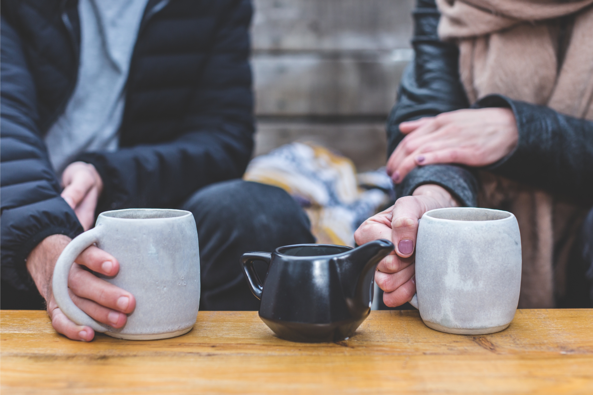 Two people sat on a bench outside with a mug in their hands and a black teapot in the middle.