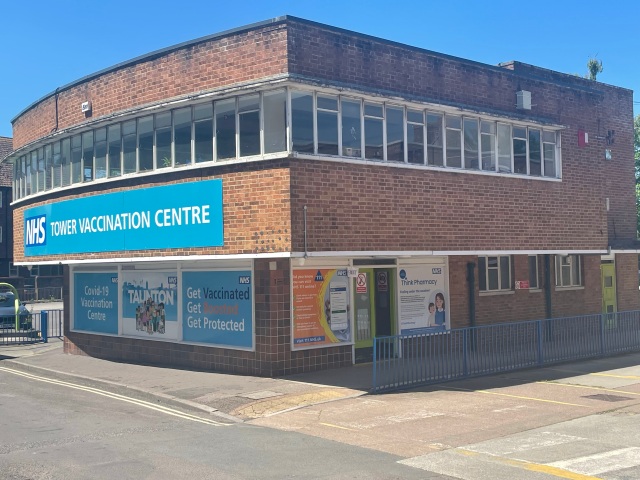Tower vaccination centre in Taunton. A red brick, flat roof building with a row of white wooden windows and a large, blue 'NHS Tower Vaccination Centre' sign.